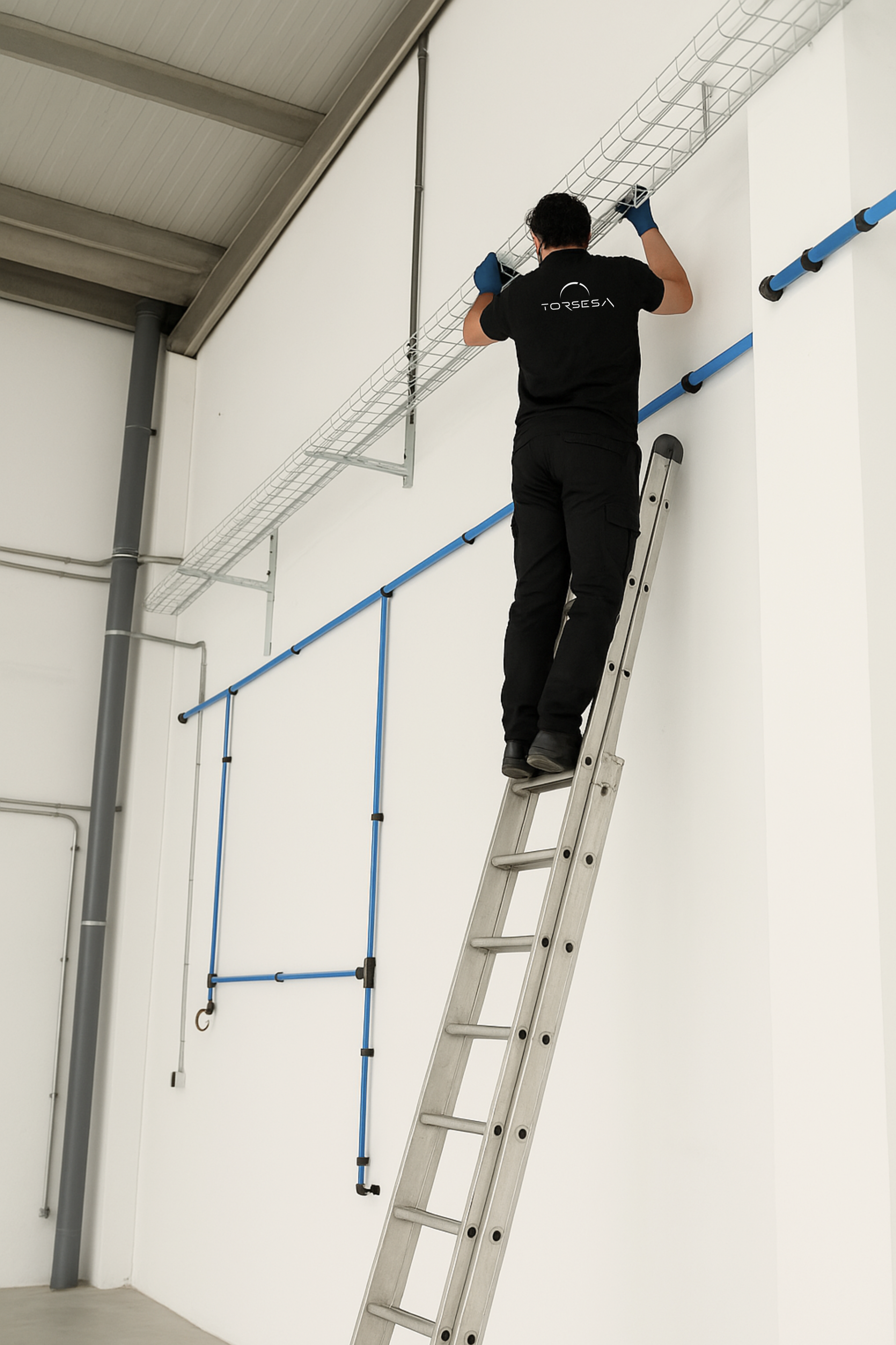 Torsesa, técnico en escalera, instalación de canaletas, red eléctrica, trabajo en altura, nave industrial, operario con guantes, montaje de infraestructura, seguridad laboral, canaleta metálica, cableado estructurado, precisión técnica, espacio limpio y ordenado.